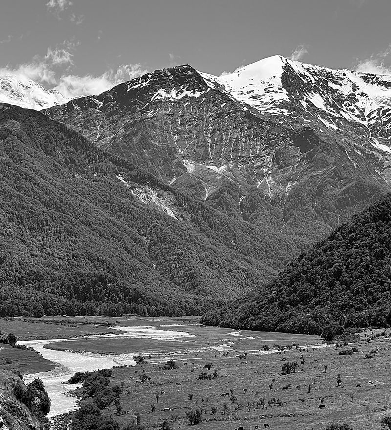 A river winding through a valley below snow-capped mountains, in black and white.